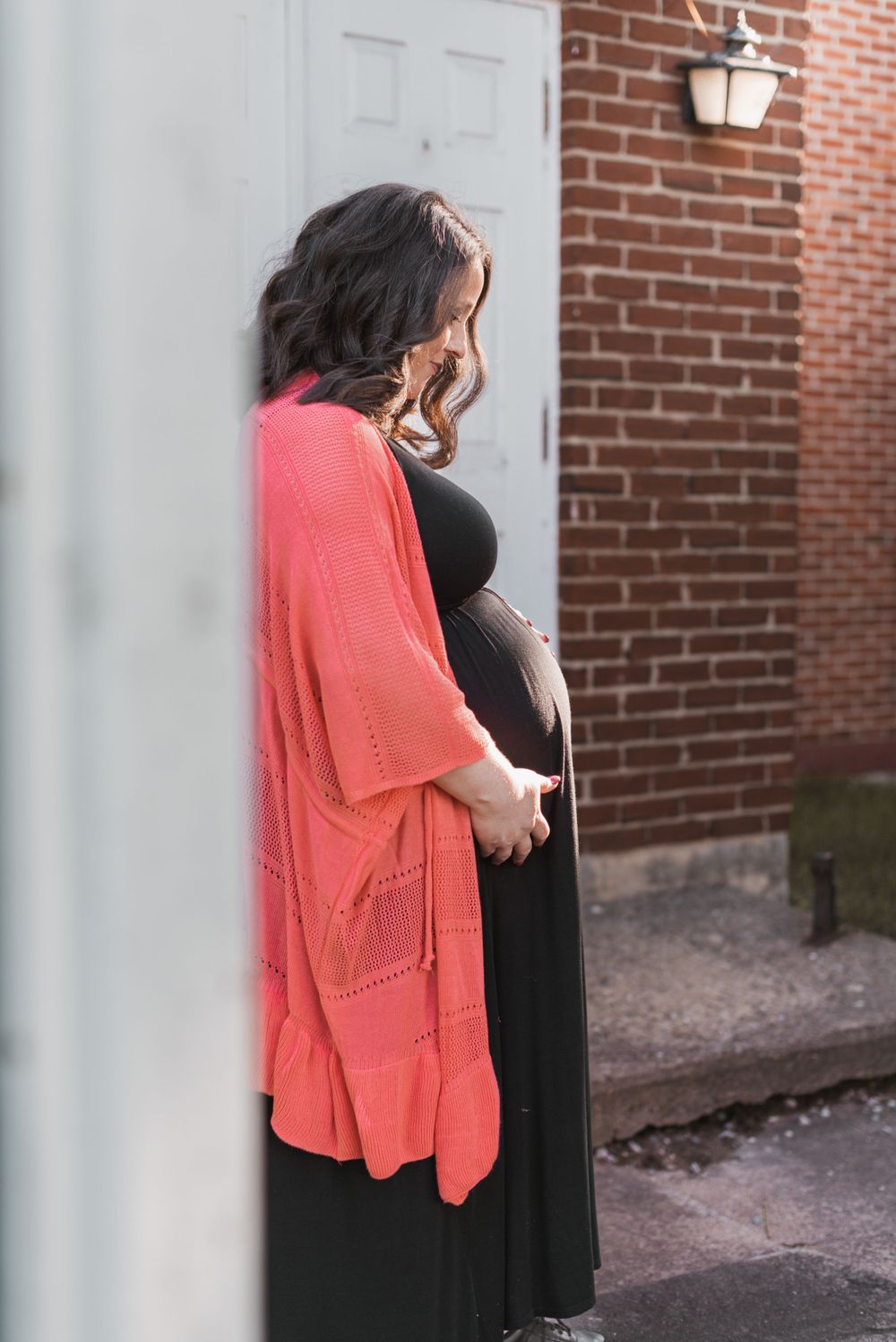 Figure wearing black dress and coral cardigan stands against brick wall.