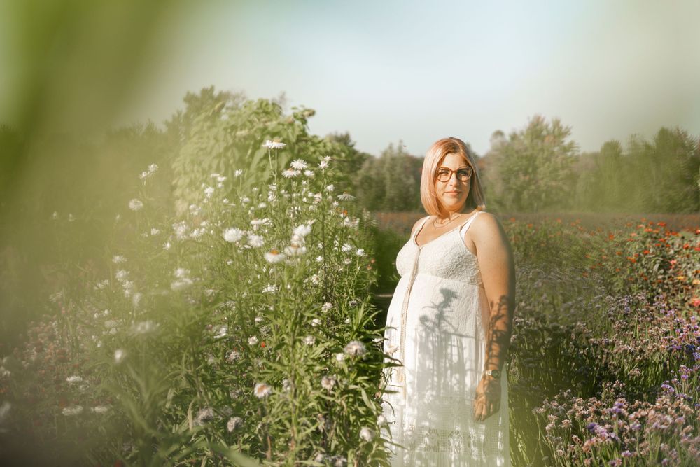 Maternity portrait of a woman in white dress standing among wildflowers in soft lighting.