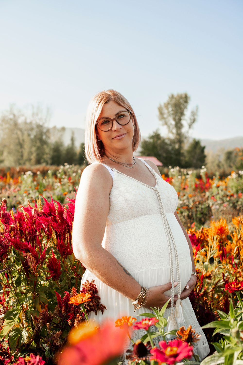 Expectant mother in a white dress poses in a vibrant flower field during golden hour.