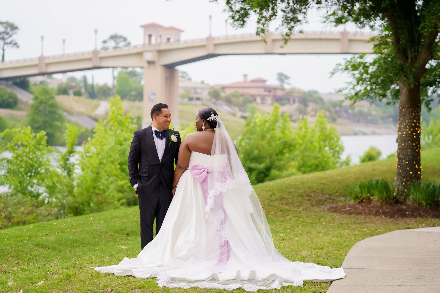 Bride and groom pose on path near bridge with bride wearing gown with pink accents.