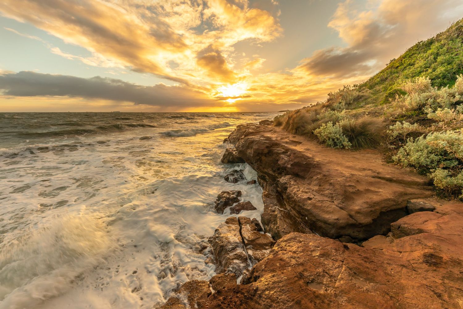 Golden sunset illuminates rocky coastal cliffs with crashing waves below.
