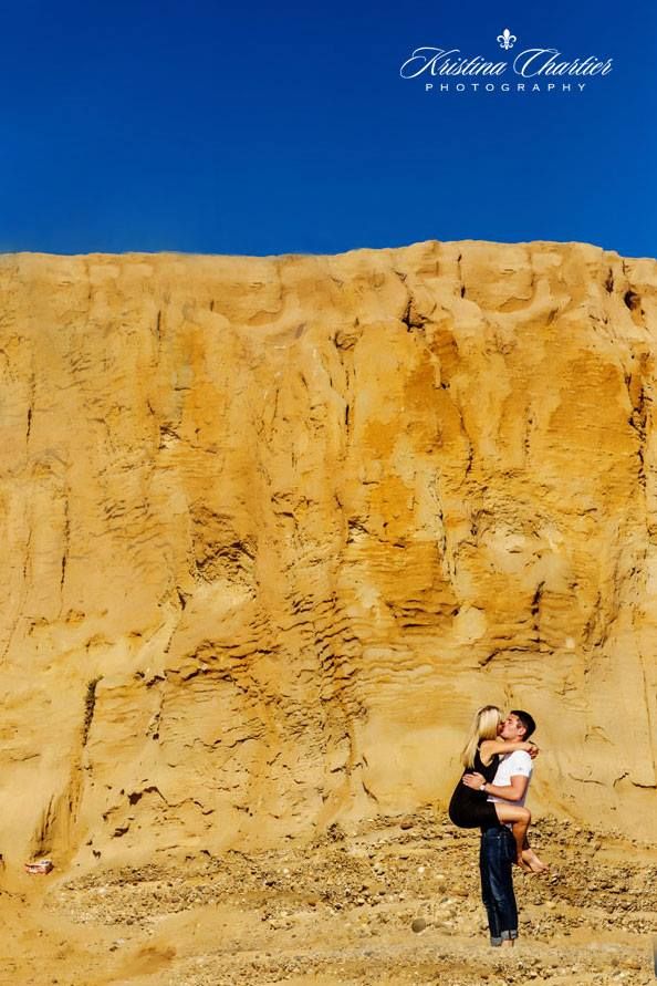 A couple embraces against a dramatic golden cliff face under bright blue sky.