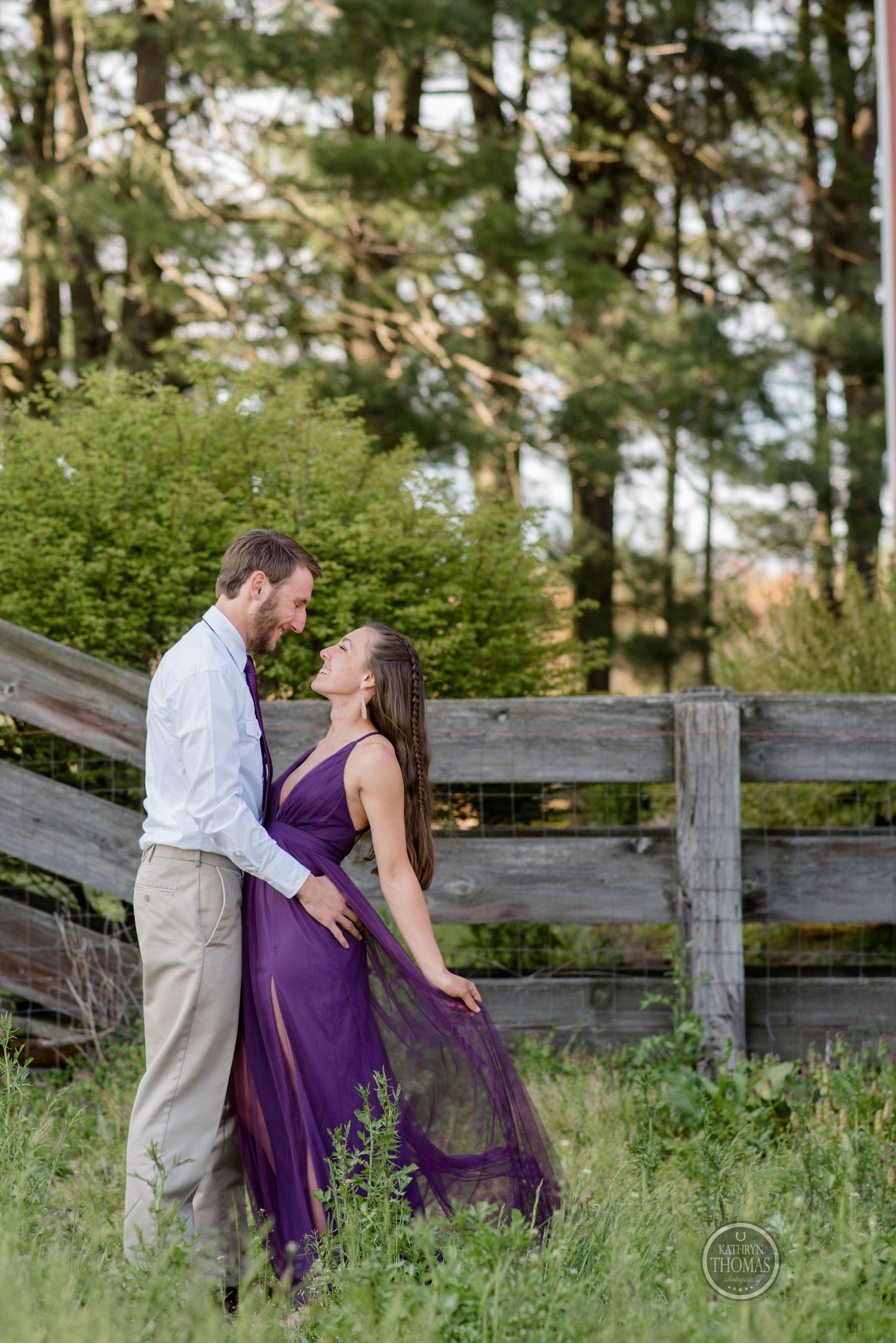 Romantic couple pose by wooden fence in purple dress and white shirt amid lush greenery.