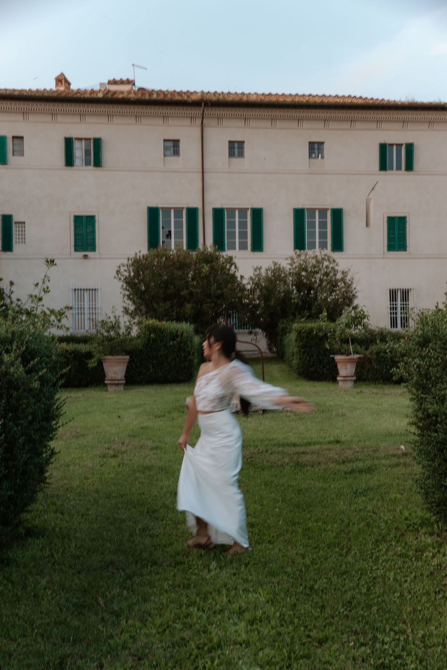 A person in a white dress spins in a garden in front of an Italian villa with green shutters.