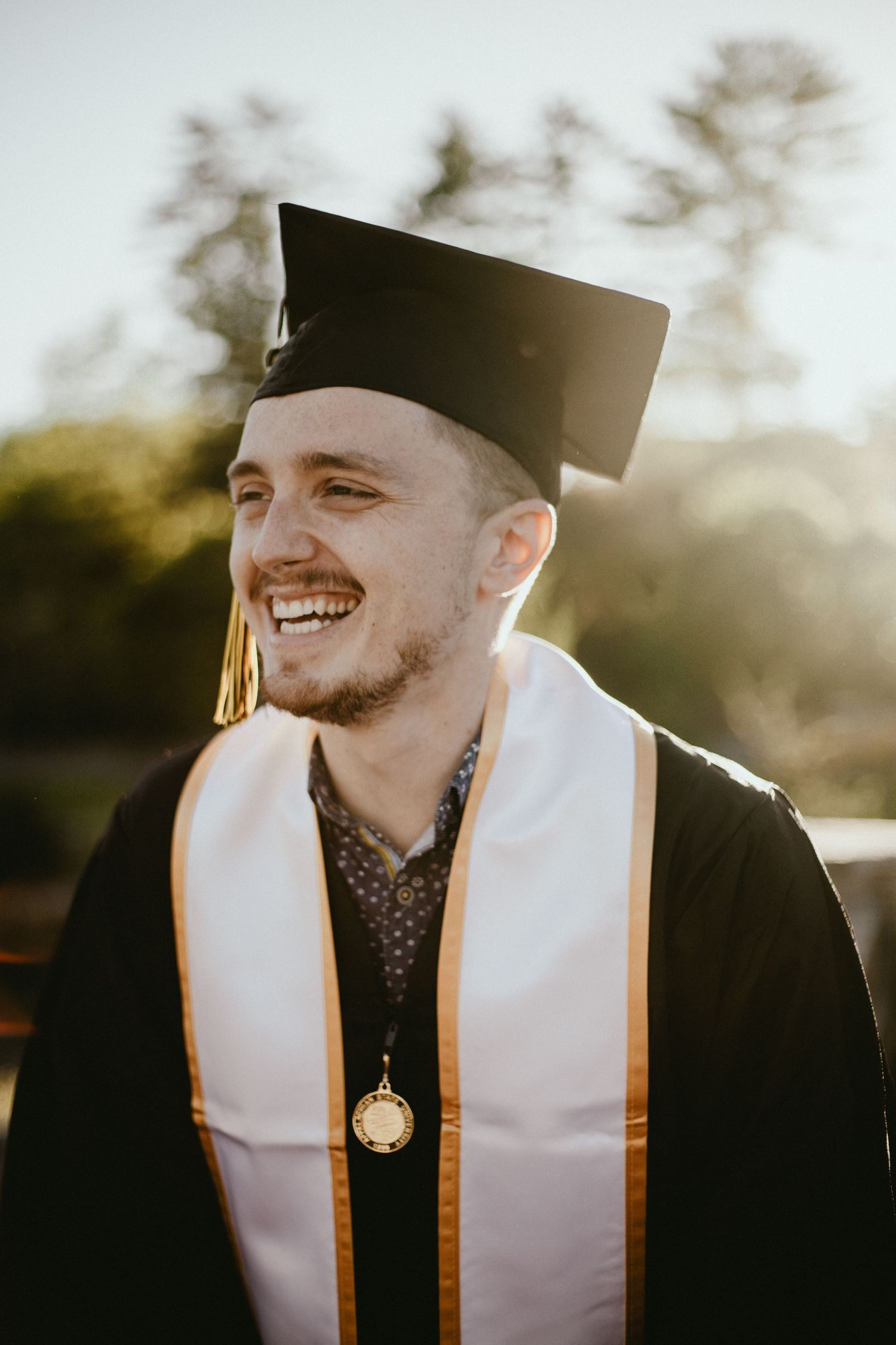 Graduate in black cap and gown with gold stole smiling in warm sunlight.