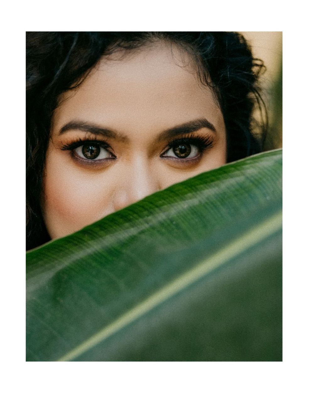 Close-up artistic portrait showing eyes peeking over a large green tropical leaf.