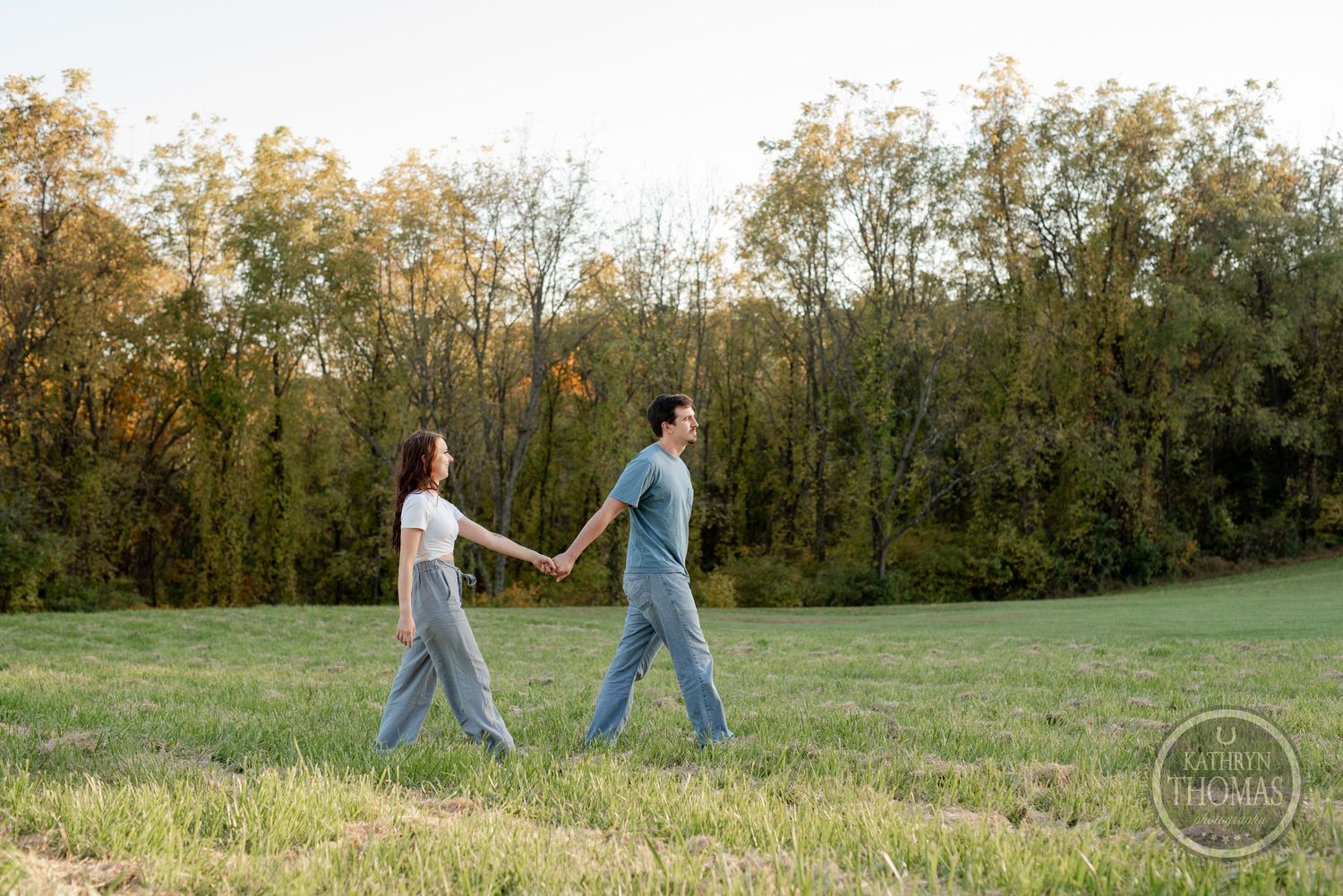 A couple holds hands while walking through a grassy field at sunset.