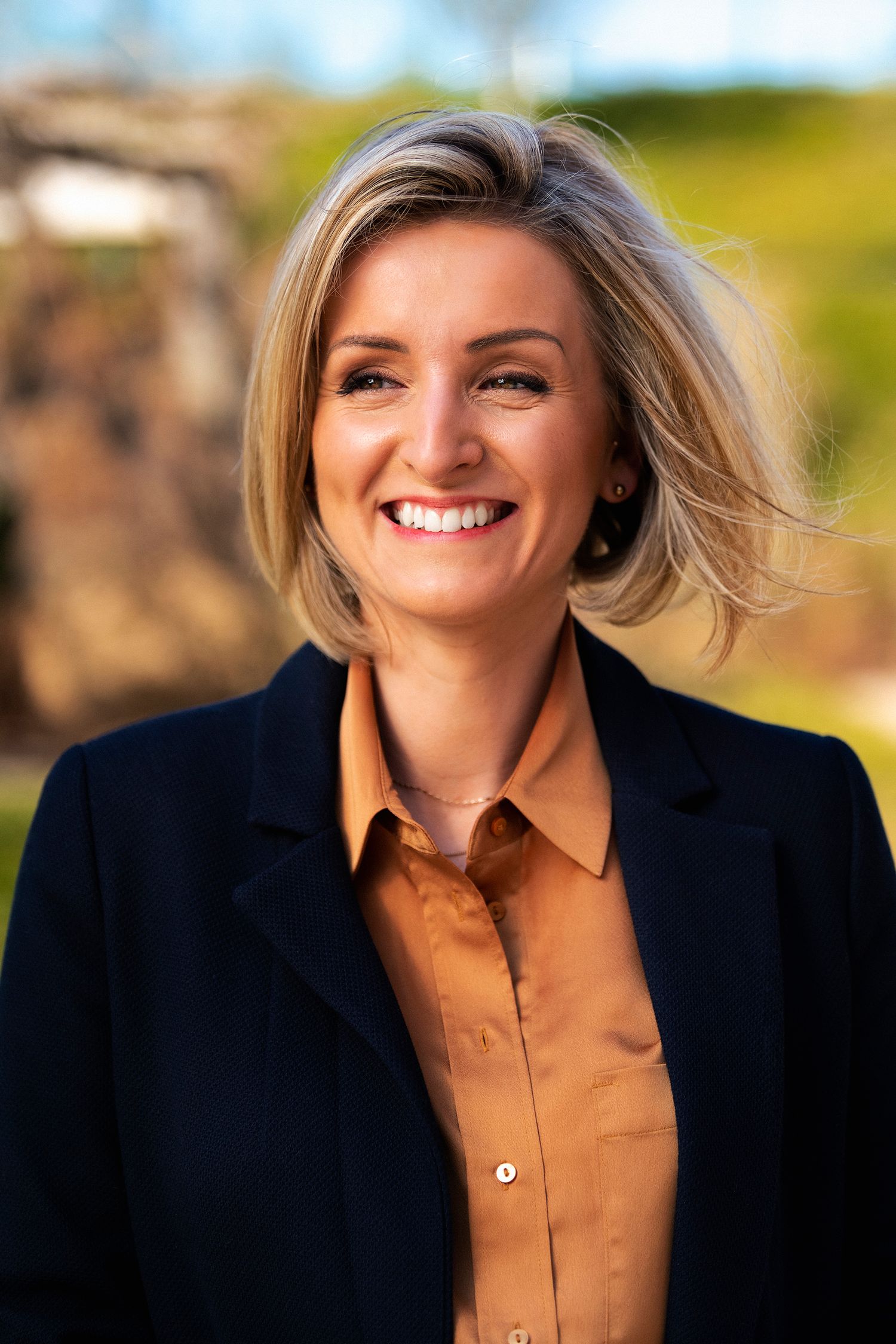 Professional headshot of person in navy blazer and brown shirt against blurred outdoor background.