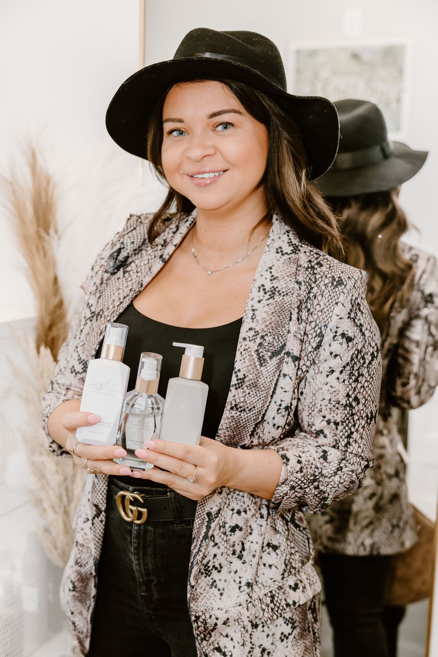A stylist in black hat and snakeskin blazer holding hair products in a salon setting.