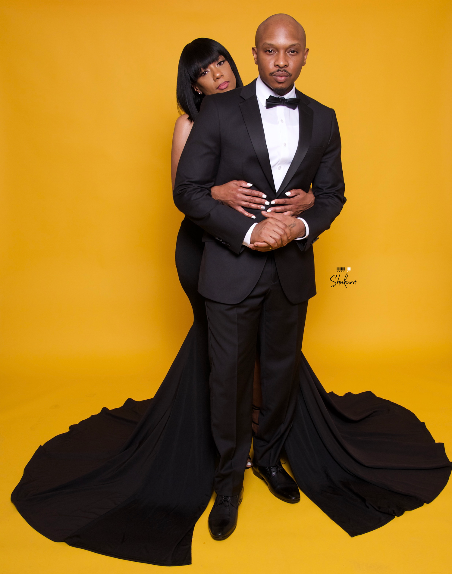 Glamorous couple in formal black attire embrace in intimate pose against yellow backdrop.