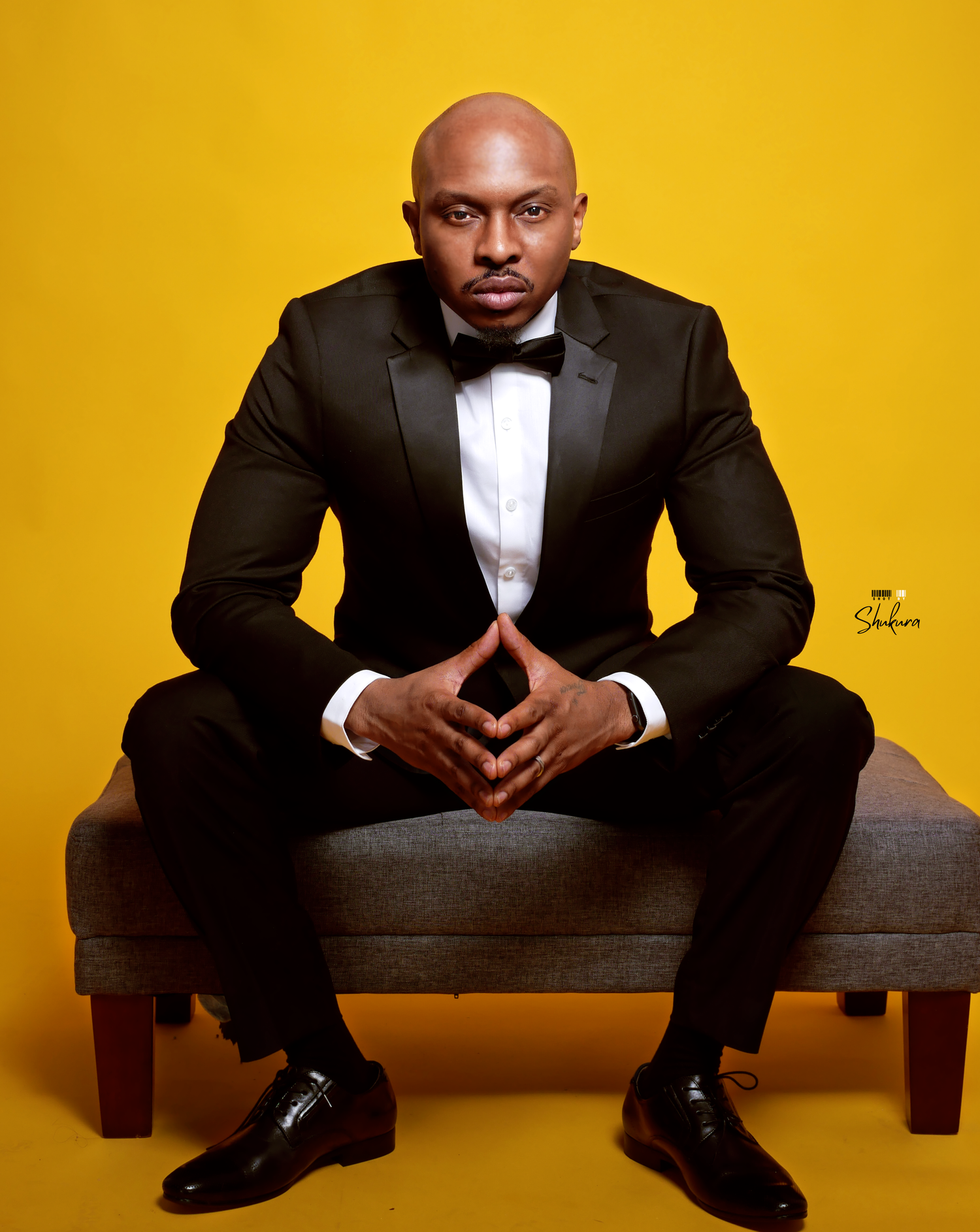 Sharp-dressed man in tuxedo seated on bench with clasped hands against yellow backdrop.