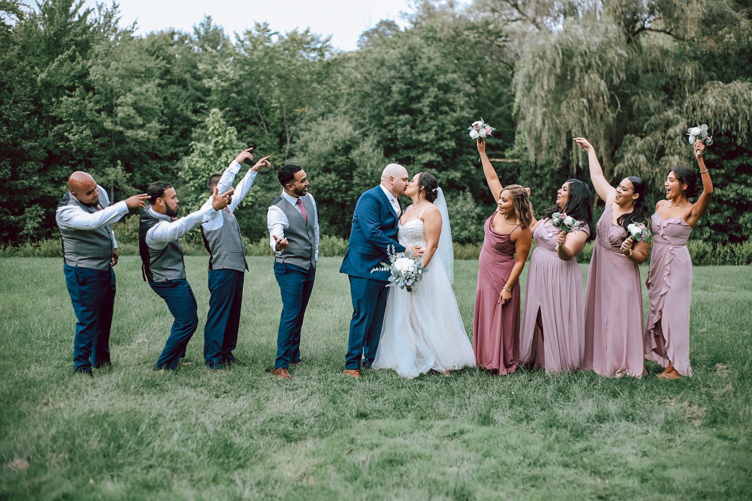 Wedding party poses playfully in a line on green grass wearing pink and gray formal attire.