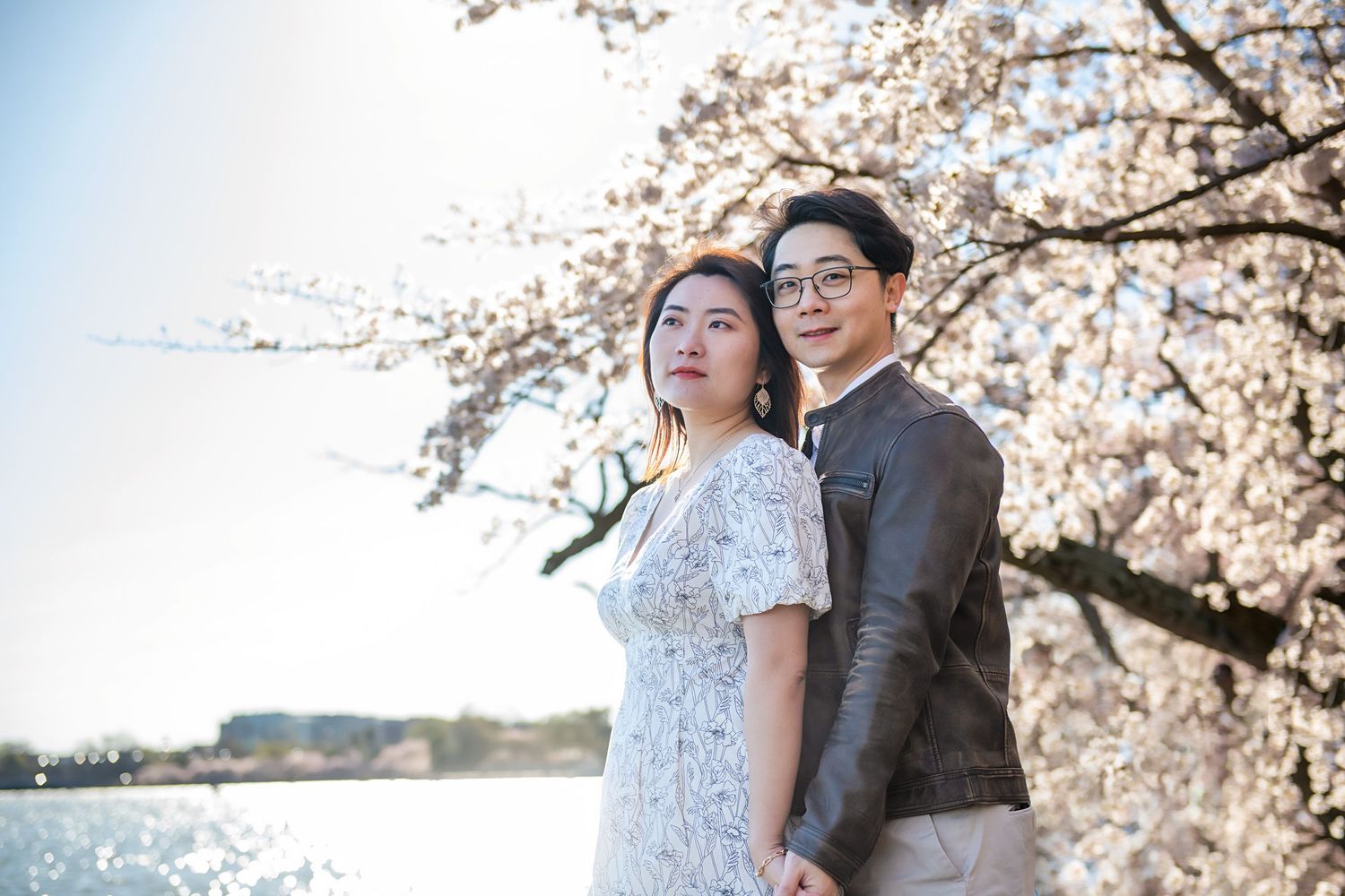 Couple in formal attire pose together under blooming cherry blossoms by waterfront.