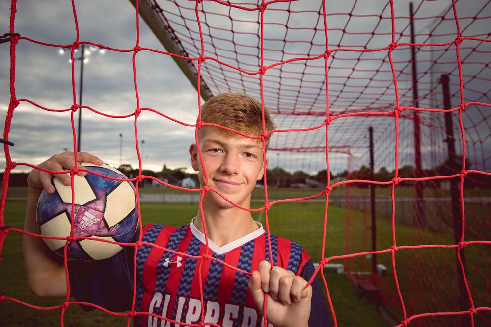 Soccer player holding ball and smiling through red goal netting on a sports field.