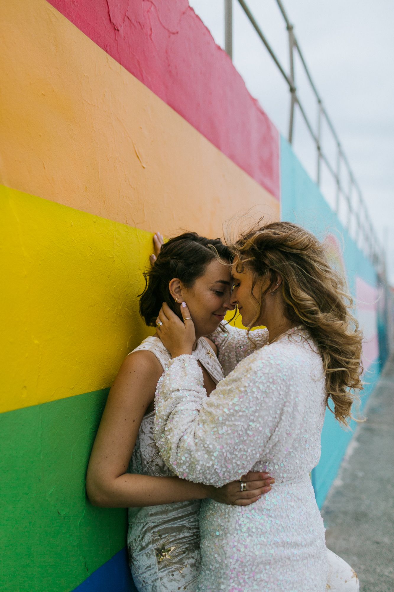 Two brides embrace intimately against a vibrant rainbow pride mural wall.