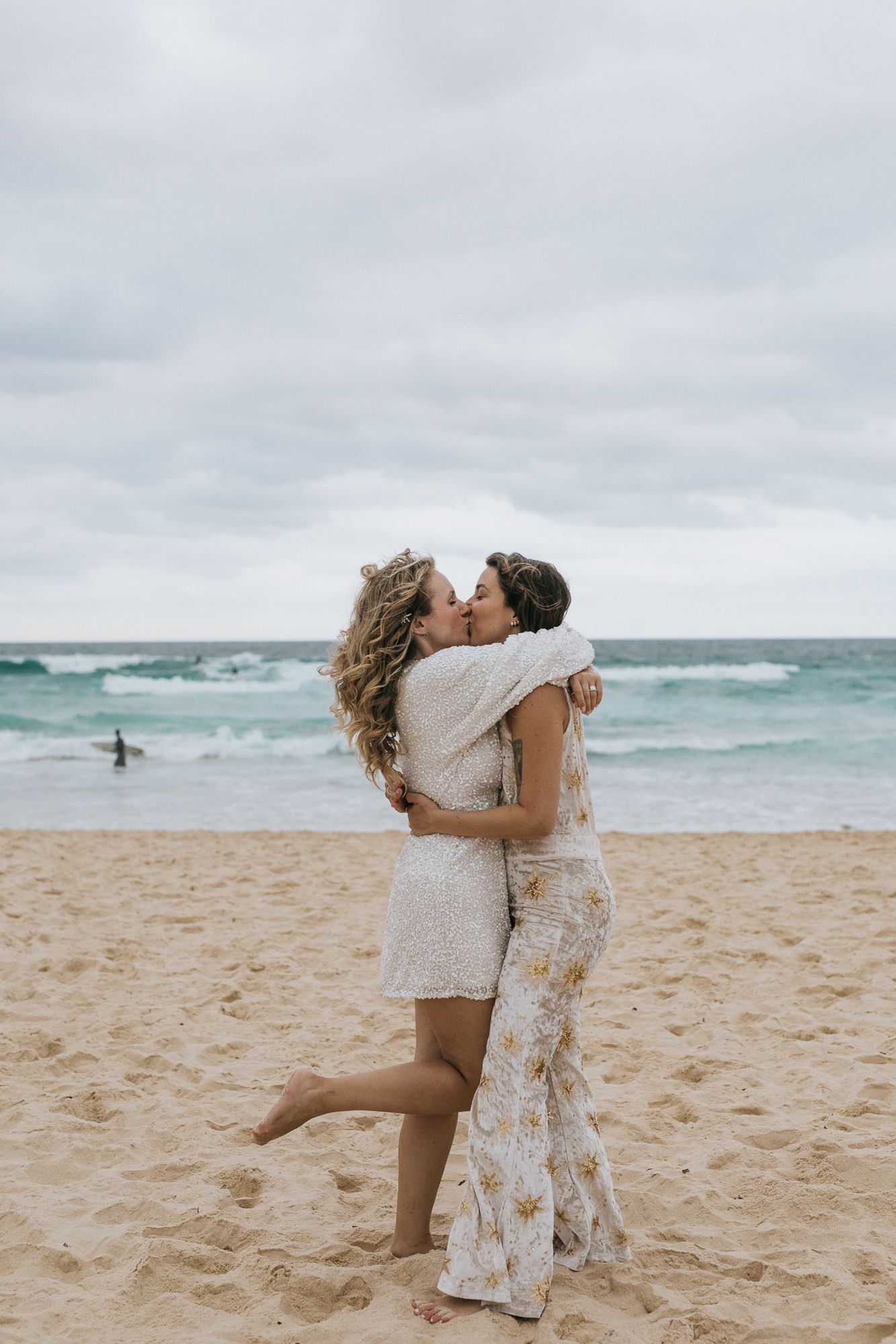 Two brides sharing romantic kiss on sandy beach with ocean backdrop.