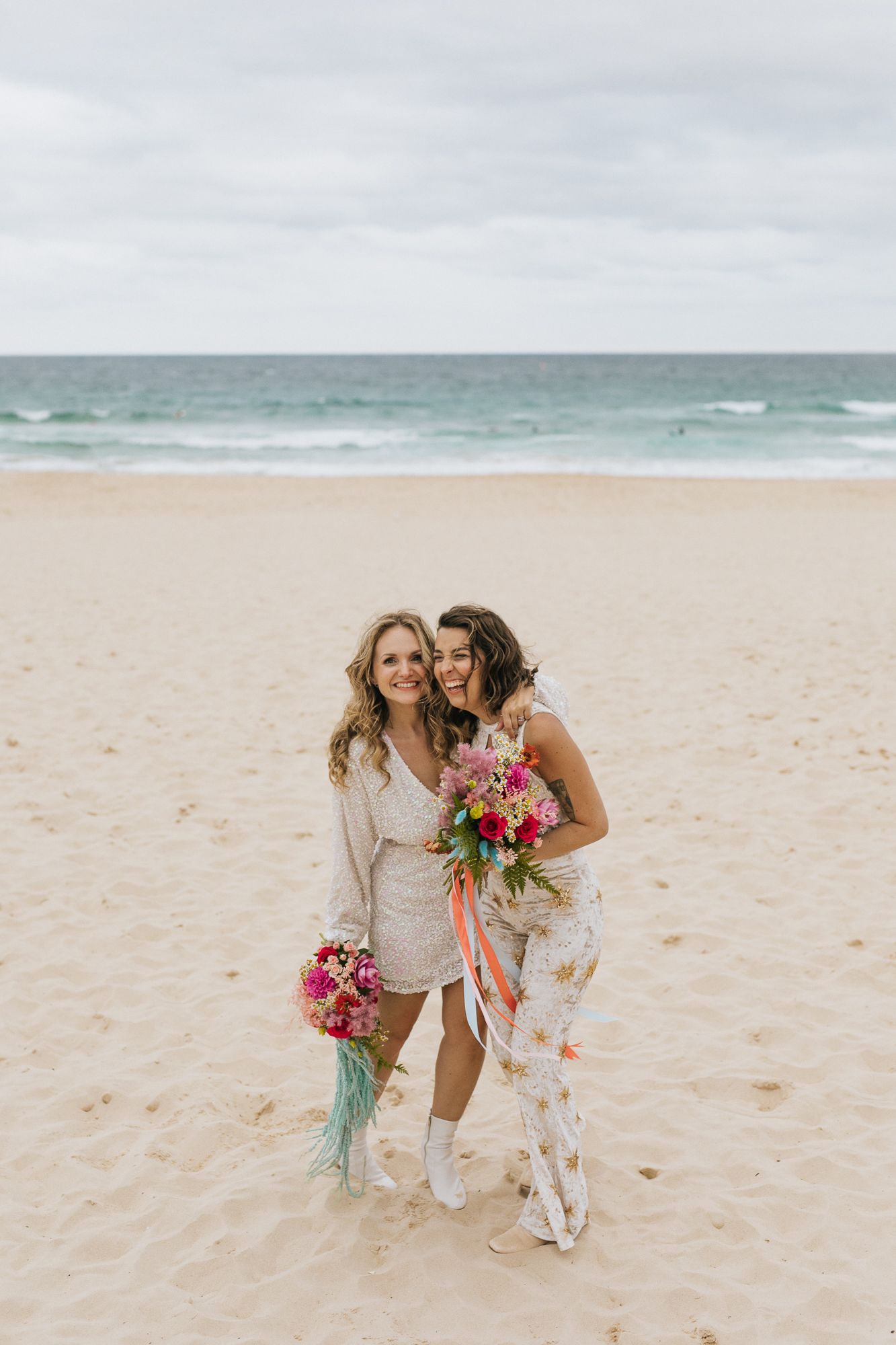 Two brides embracing on sandy beach holding bright colorful wedding bouquets.