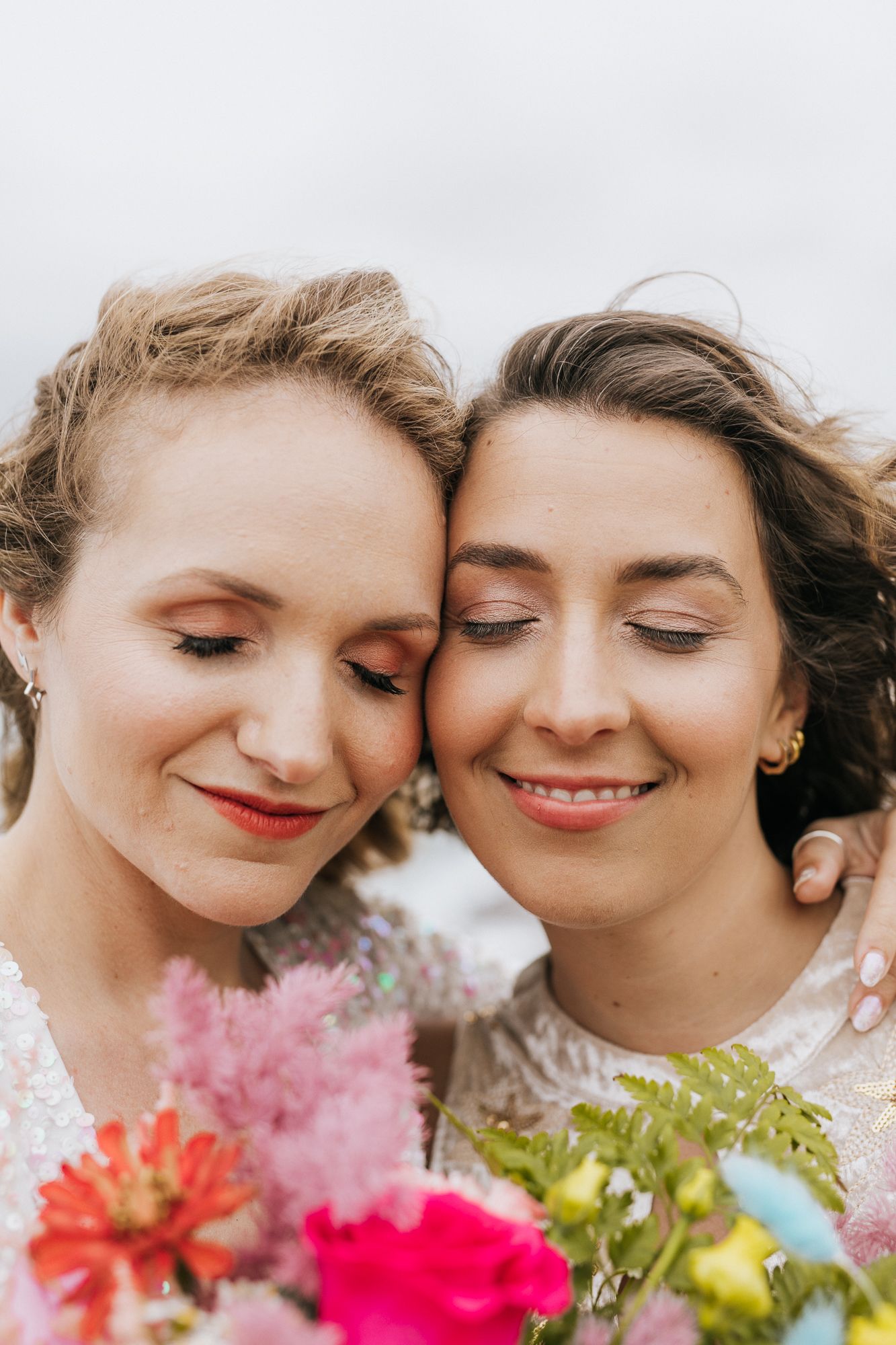 Two brides sharing tender moment with eyes closed holding vibrant floral bouquets.