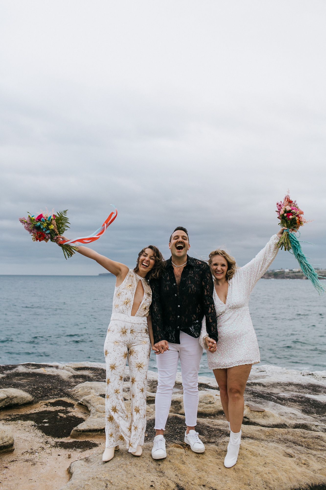 Two brides in white holding colorful bouquets celebrate with officiant on rocky ocean shore.