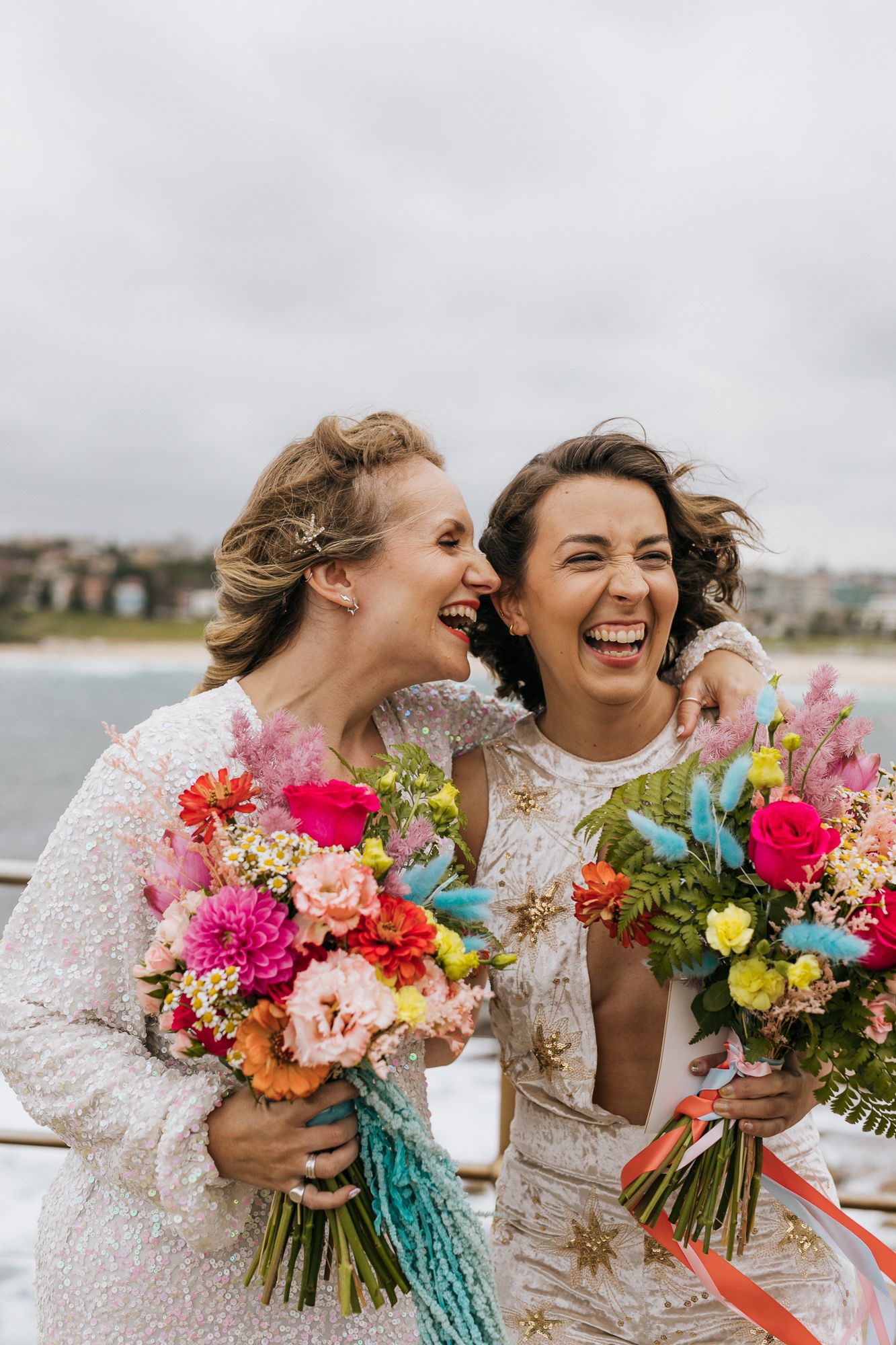 Two brides laughing joyfully holding colorful bouquets at Bondi Beach wedding.