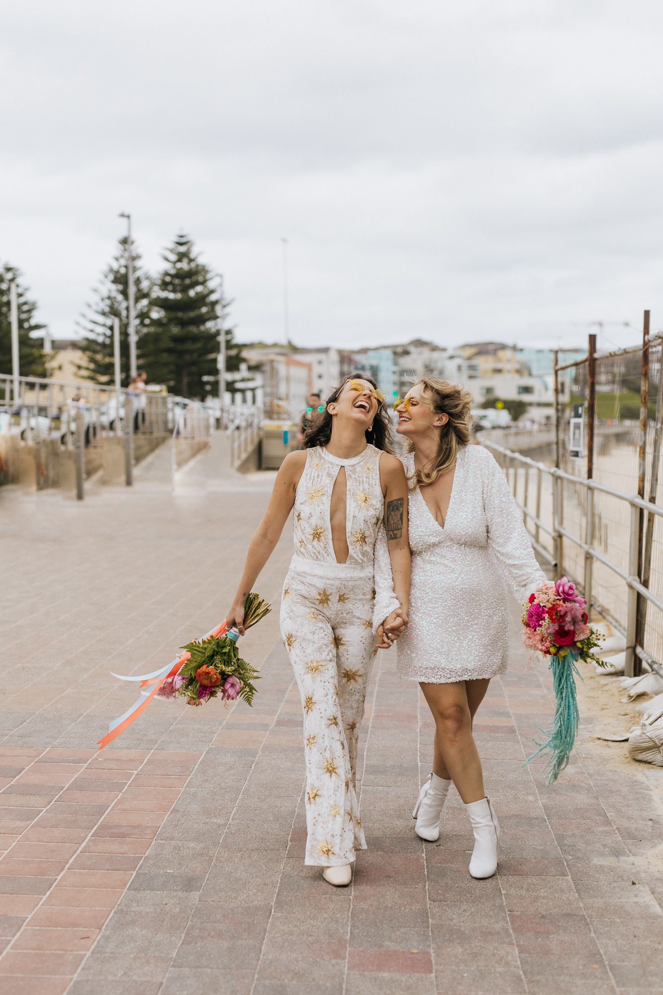 Two brides walking hand in hand laughing along Bondi Beach promenade.