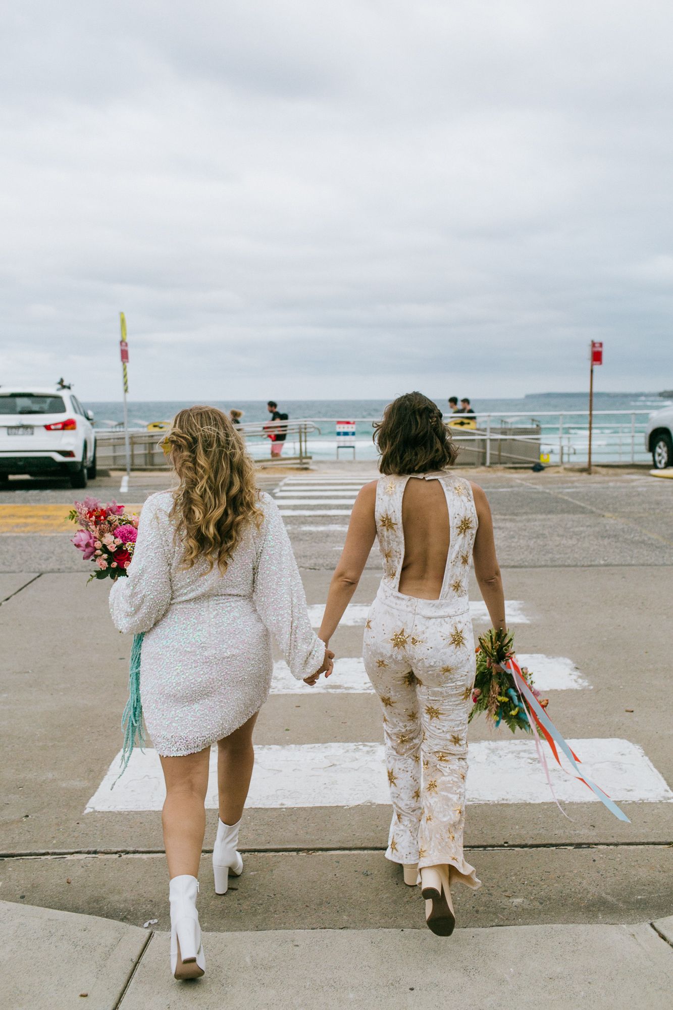 Two brides in white holding bouquets walk hand in hand toward beach on crosswalk.