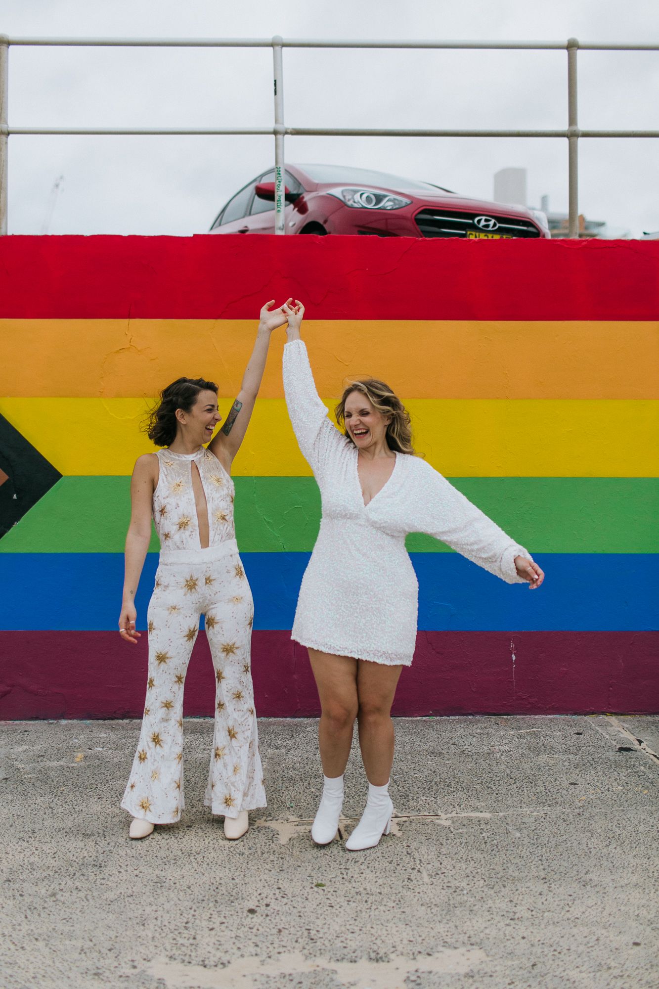 Two brides celebrate joyfully holding hands in front of a pride rainbow flag mural.