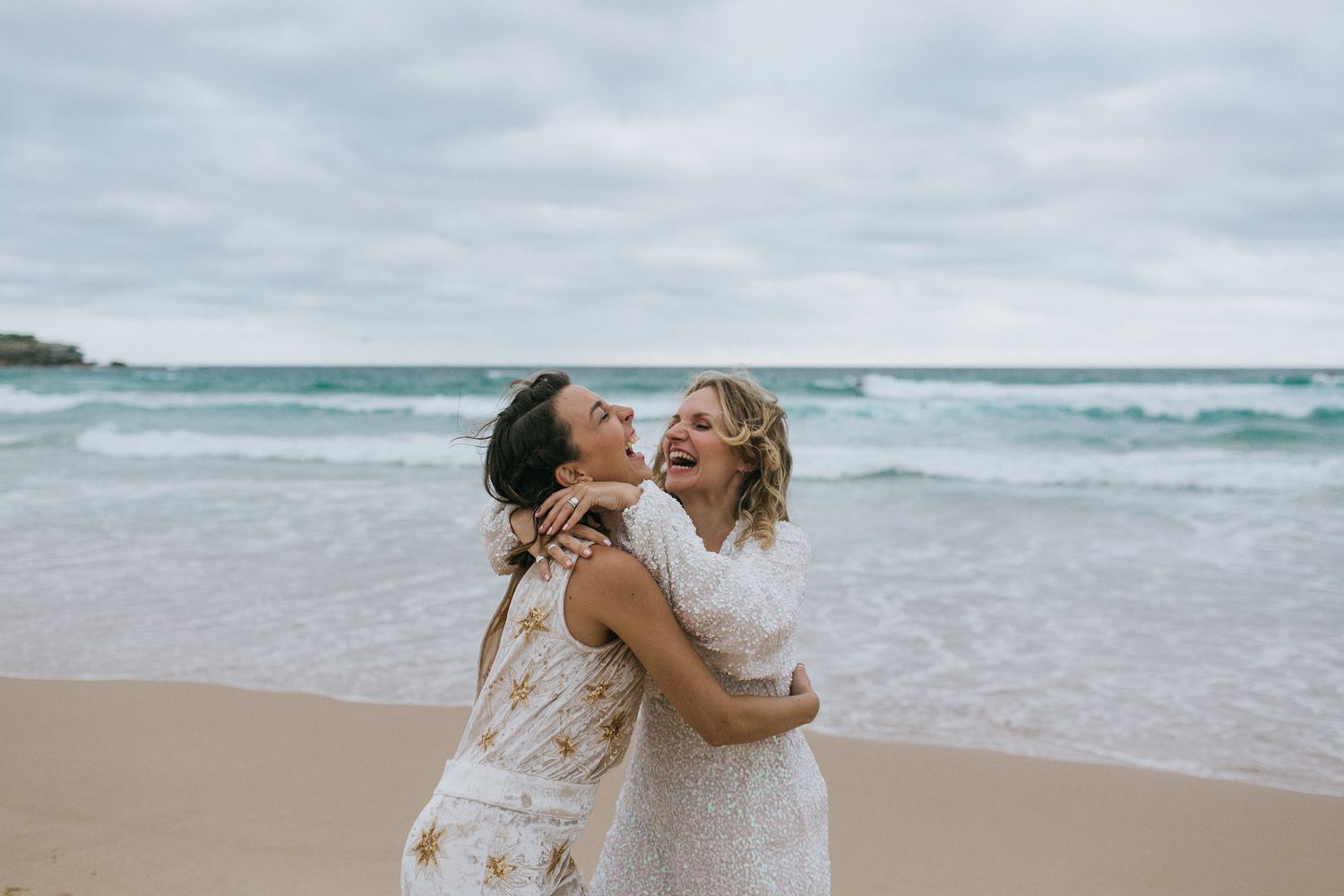Two brides laugh and embrace joyfully on a sandy beach with ocean waves behind.