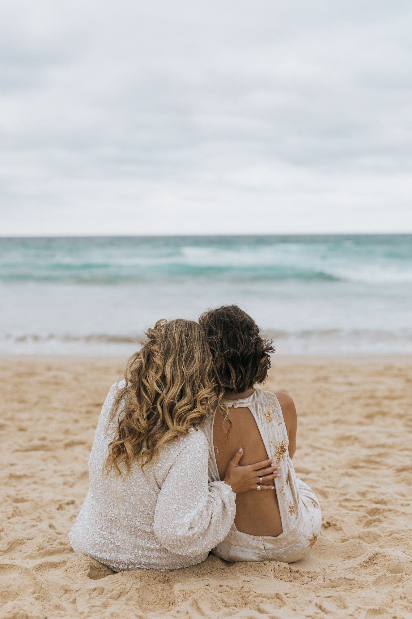 Two brides sitting together on sandy beach gazing at ocean waves.