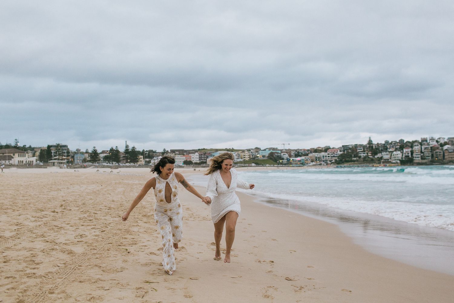 Two brides in white run hand in hand along sandy Bondi Beach shoreline.