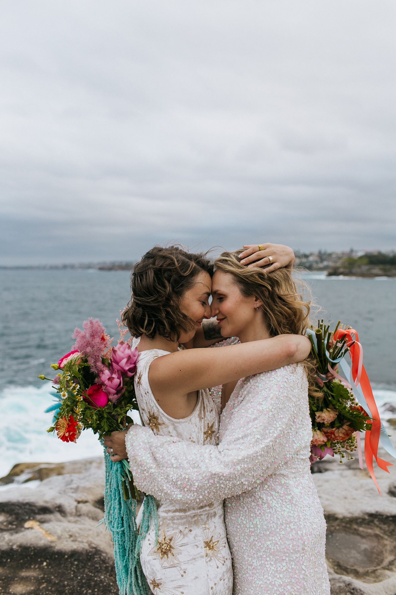 Two brides embracing forehead to forehead holding vibrant bouquets by ocean.