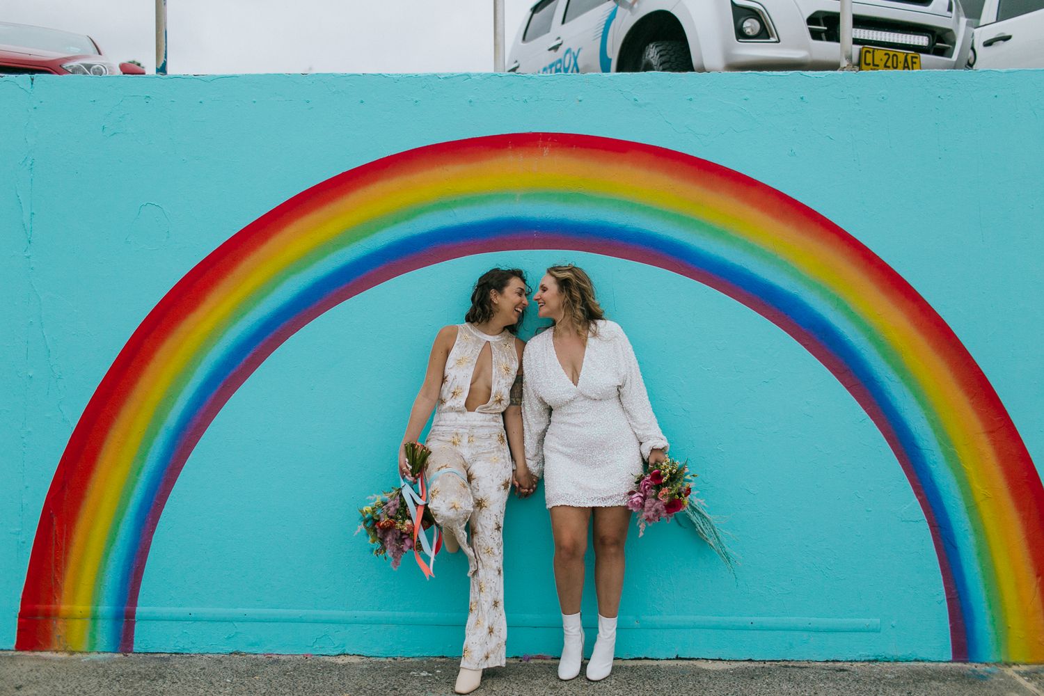 Two brides pose lovingly under a rainbow mural on a bright turquoise wall.
