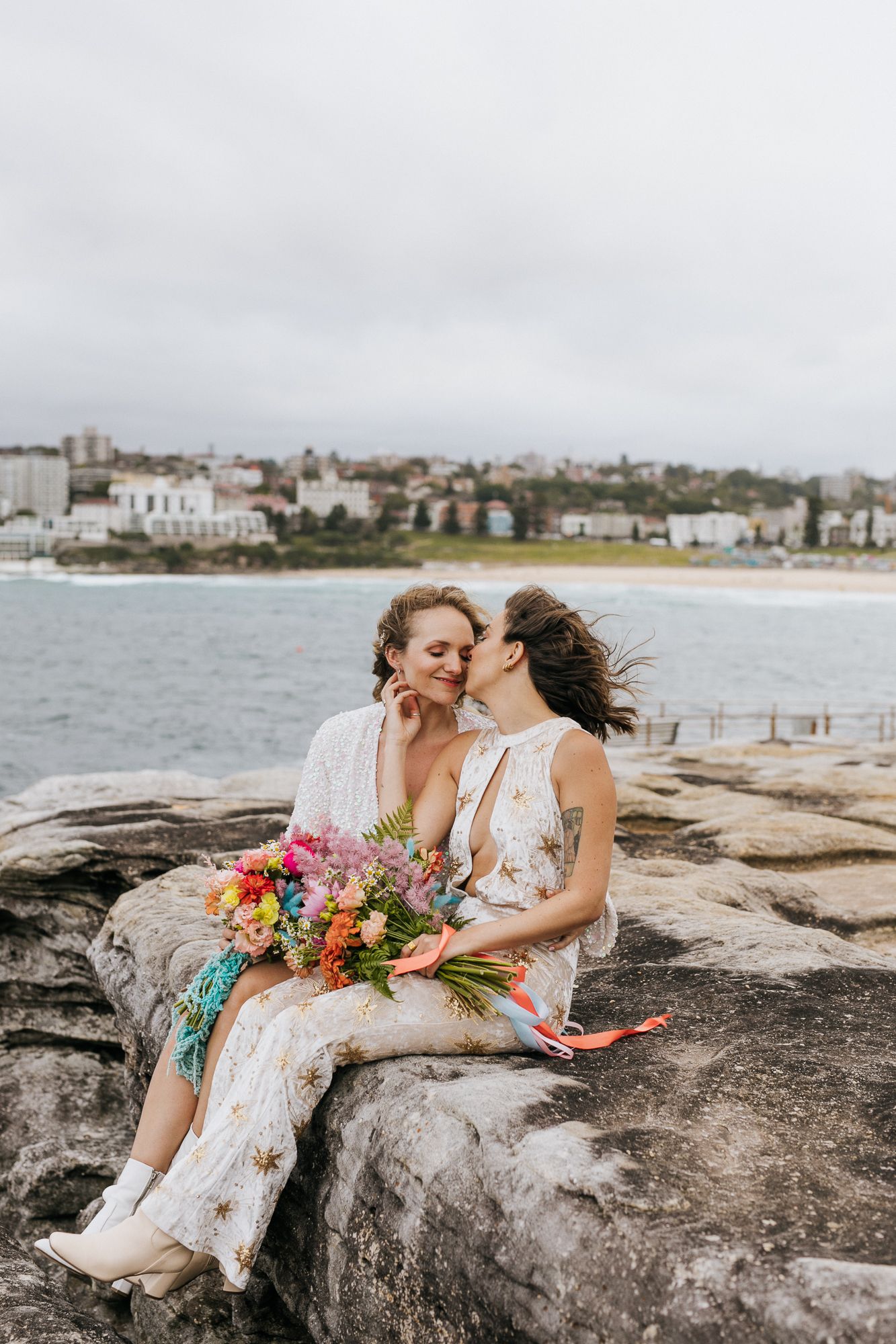 Two brides sharing kiss on rocky Bondi Beach coastline with colorful bouquet.