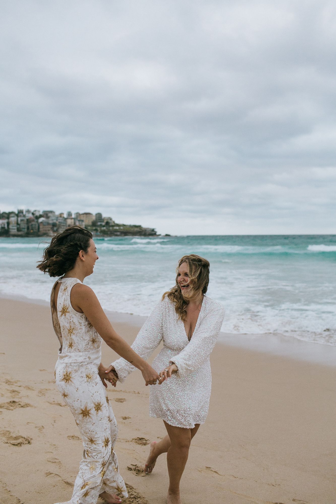 Two brides hold hands and smile at each other while walking along the beach.