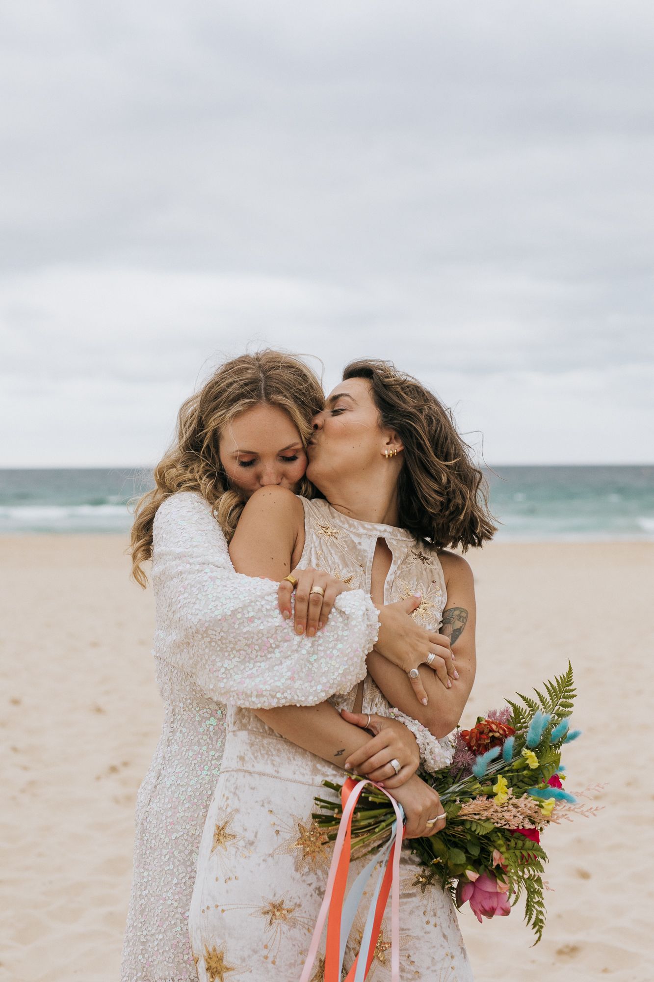 Two brides hugging and kissing on sandy beach with colorful bouquet.