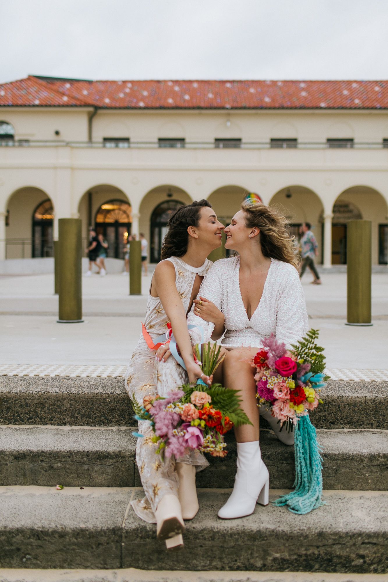 Two brides share a kiss while seated on steps holding vibrant colorful wedding bouquets.