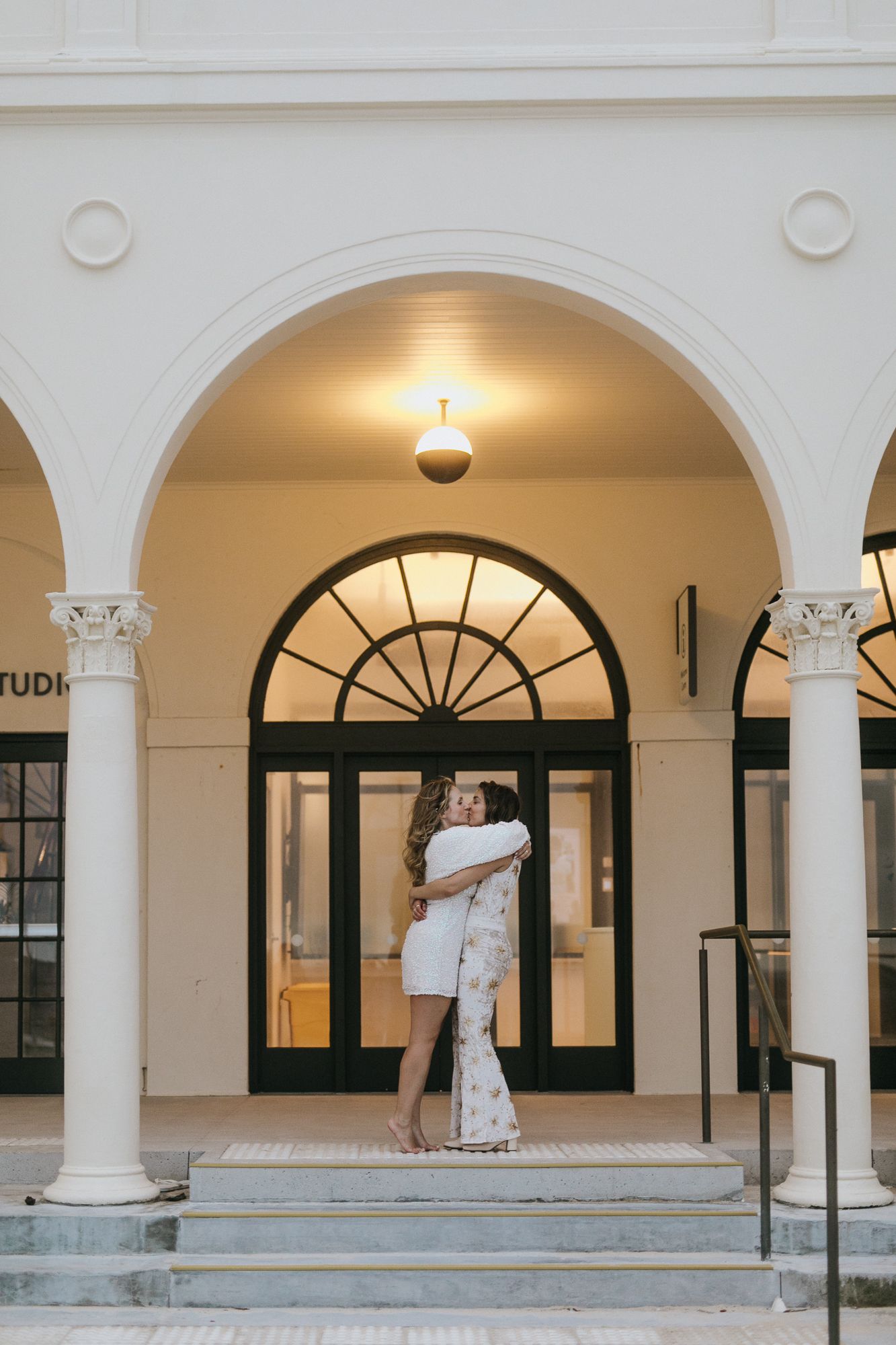 Two brides embracing under elegant white arched building entrance at wedding.