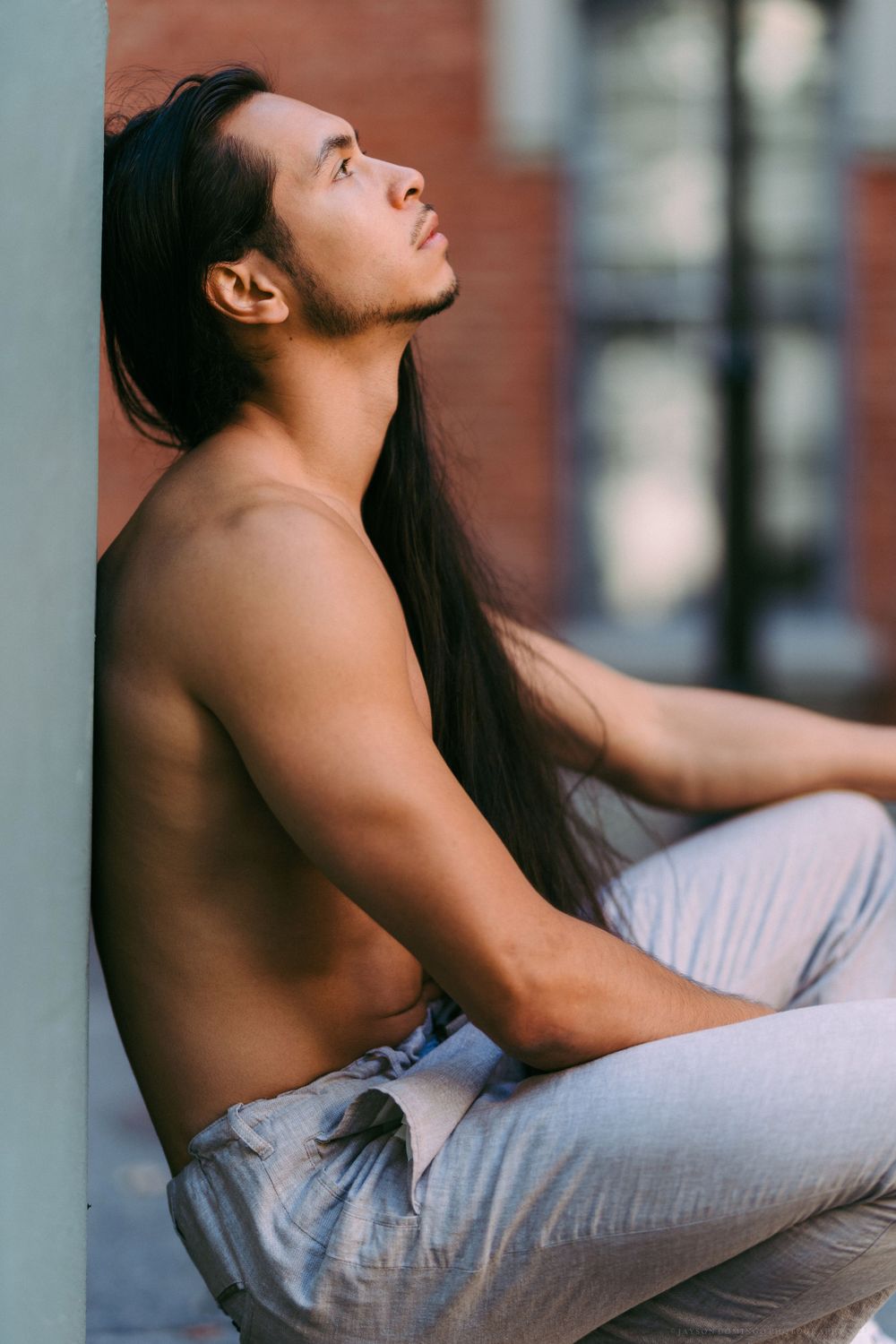 Model with long dark hair leans against wall in profile wearing light-colored pants.