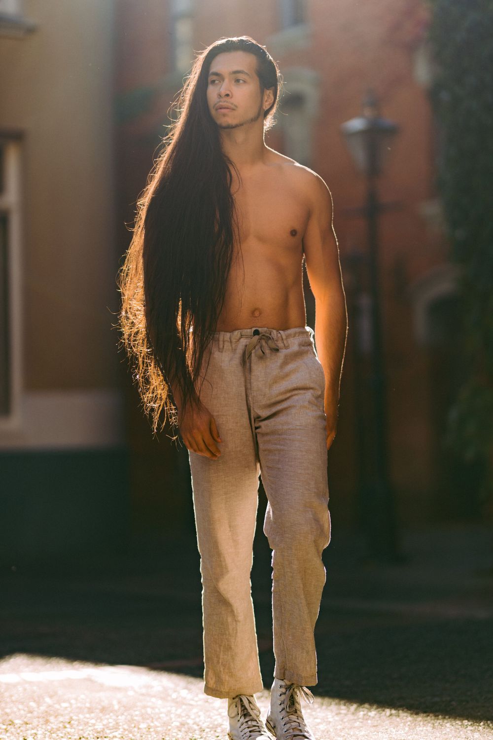 Athletic model with long dark hair poses shirtless in khaki pants against urban backdrop in golden light.