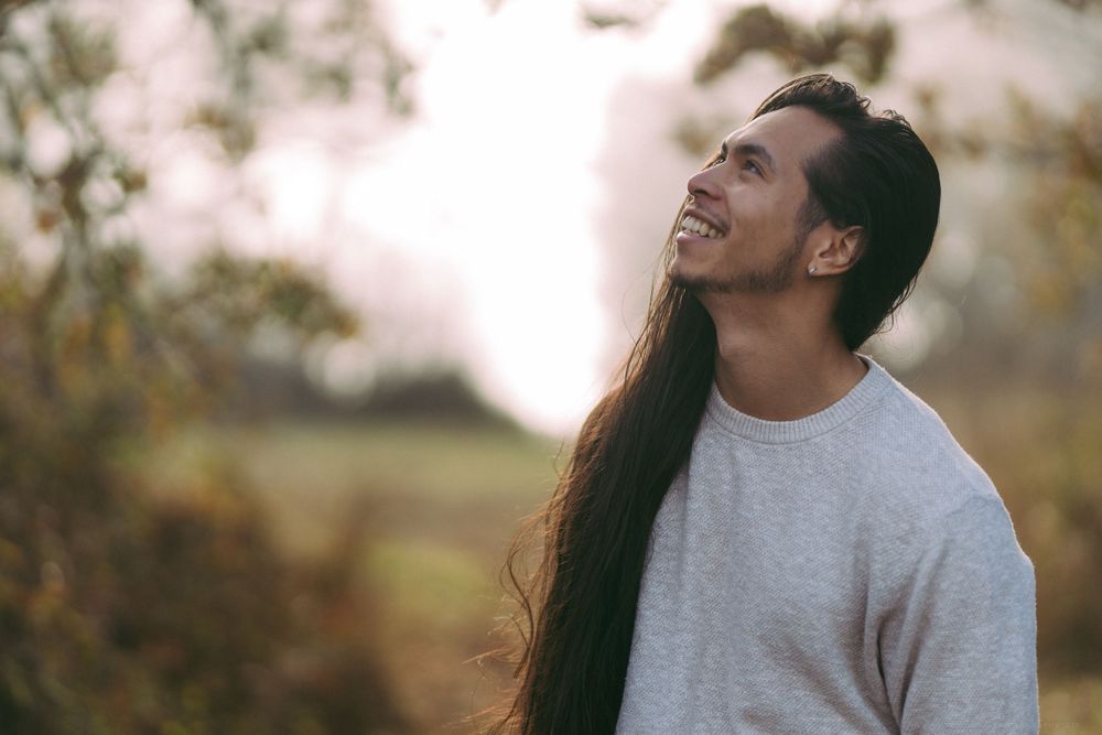 A person with long dark hair laughs joyfully while wearing a gray sweater against a soft outdoor background.