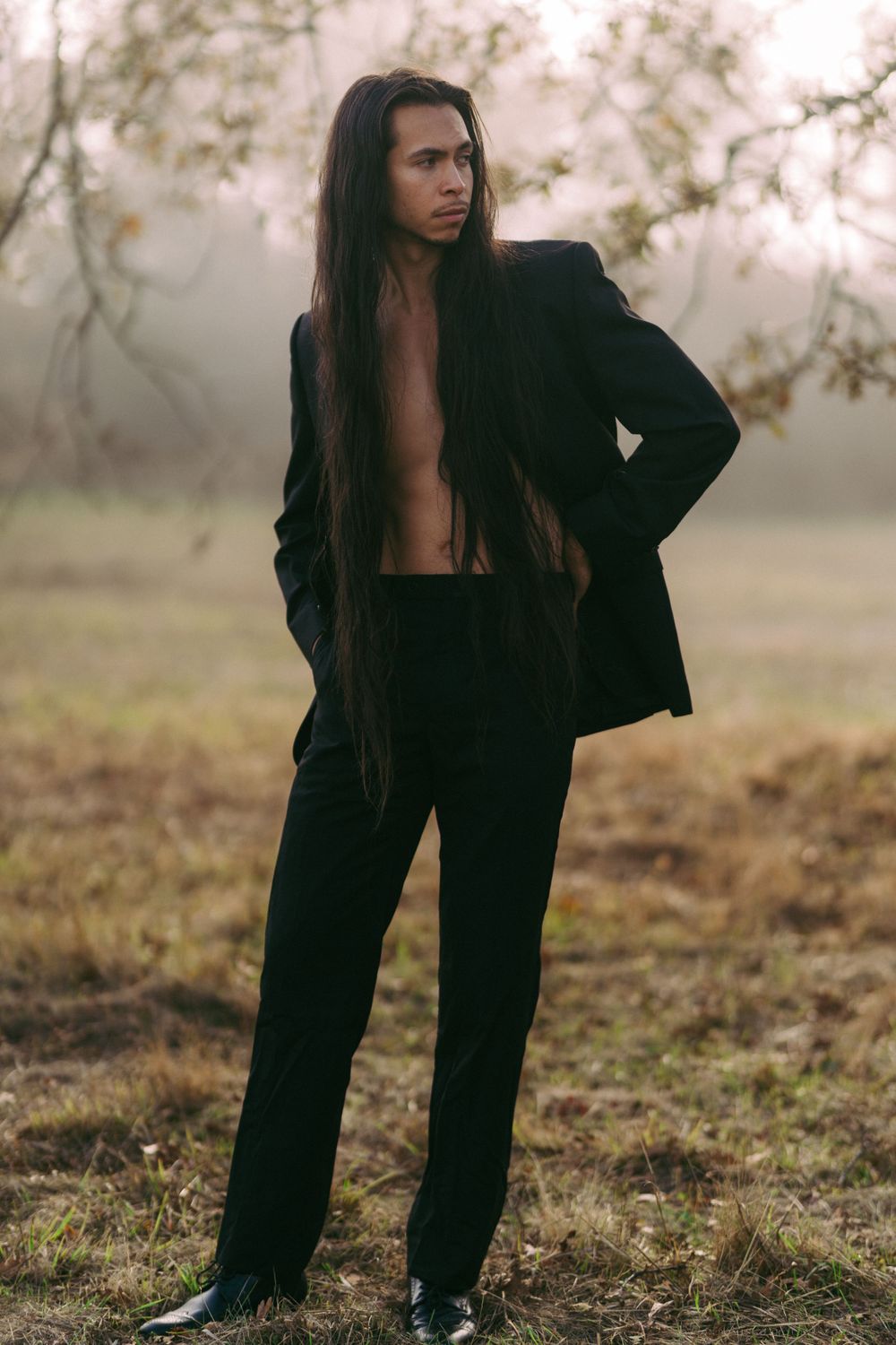 Model in open black suit poses dramatically in misty outdoor setting.