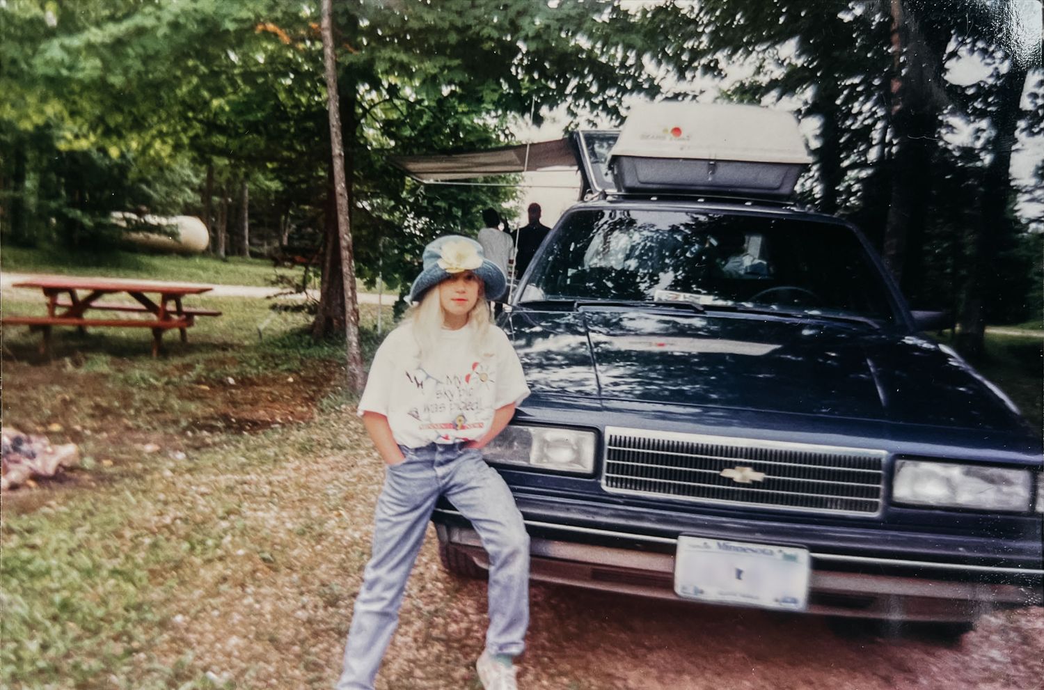 Miranda sits on the dash of an old station wagon in the early 90s. She wears a bucket hat, white T, and faded jeans.