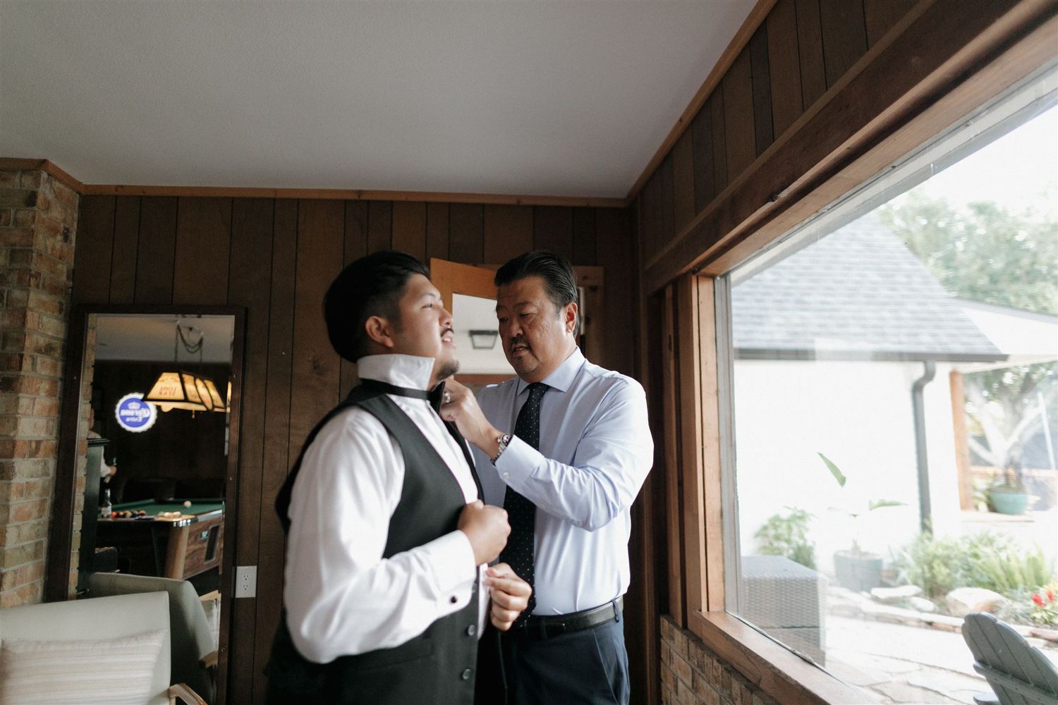 A formal tie adjustment scene in front of a window with wood paneling.