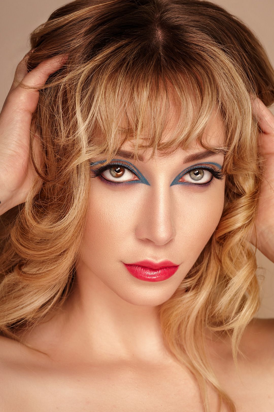 Beauty portrait with blonde wavy hair, vibrant red lips and dramatic blue eye makeup in warm lighting.