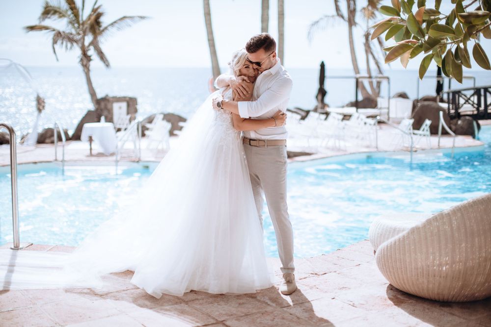 A bride and groom embracing beside a pool with a tropical backdrop, under a clear sky.