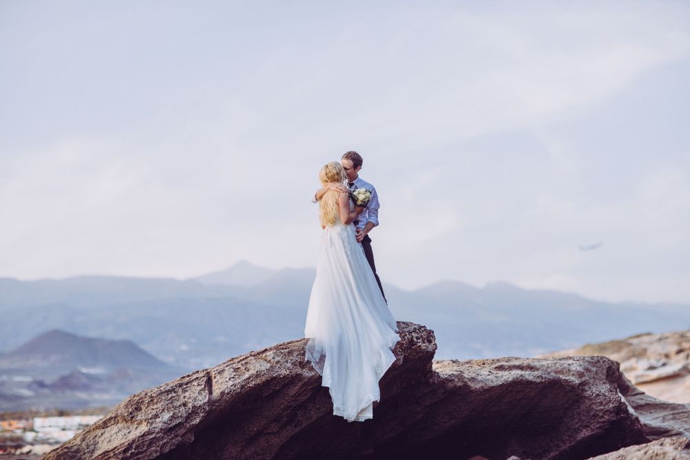 Photograph of bride and groom embrace, standing on a rock, wide shot, beach side in Tenerife South.
