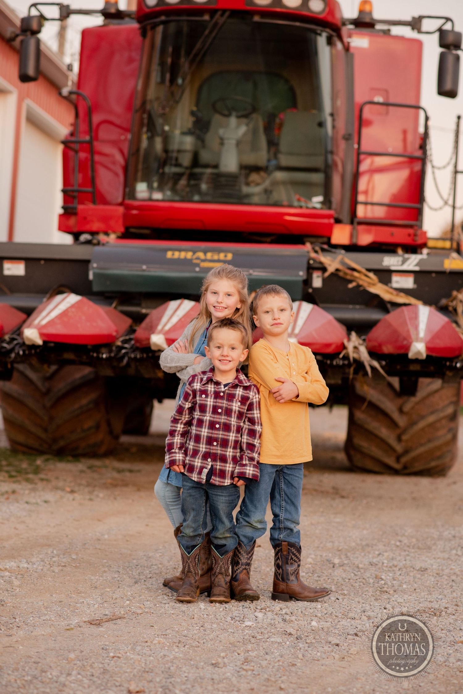 Three children posing in front of large red combine harvester on farm.