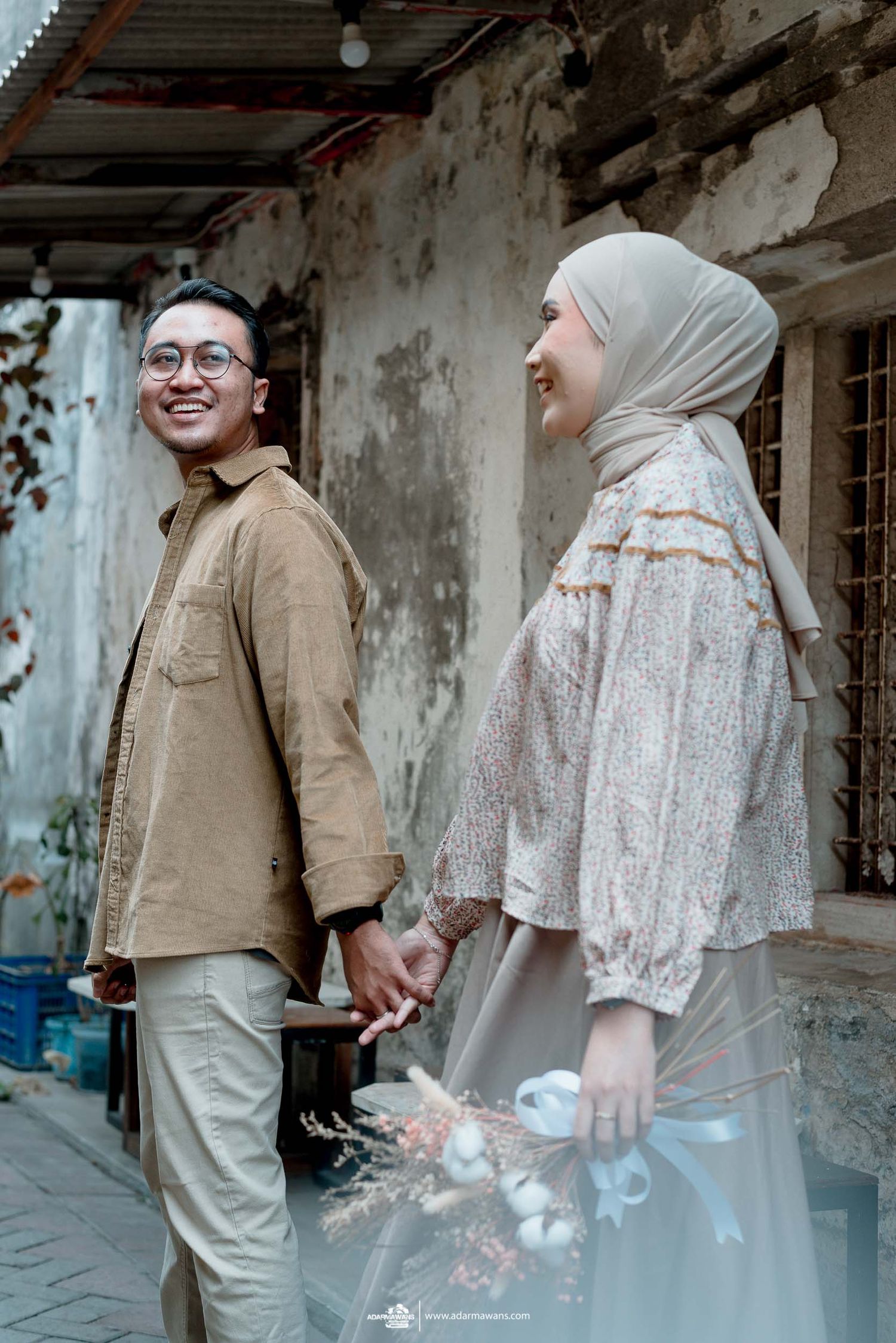 Couple holding hands and smiling while posing against weathered wall background.