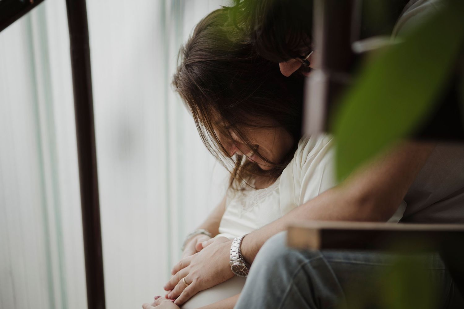 Tender indoor moment showing expectant mother looking down thoughtfully near a window.