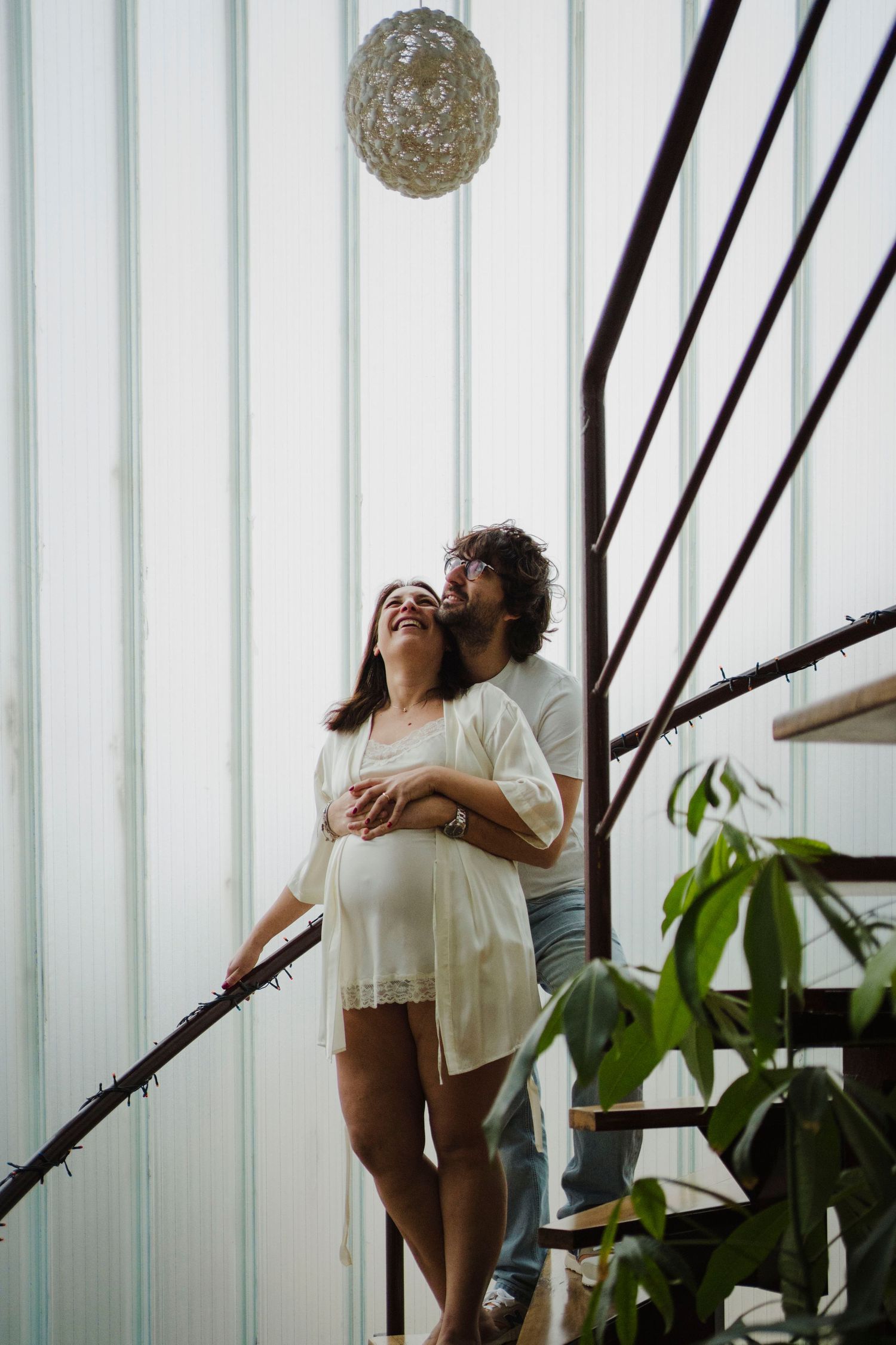 Romantic maternity moment with couple standing by stairs and decorative light fixture.